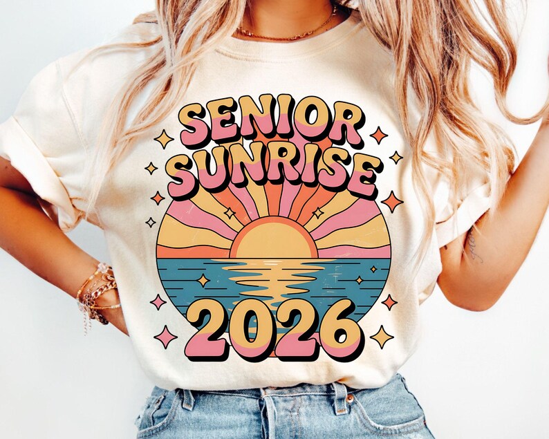 May include: Cream-colored t-shirt featuring a retro design with the words "SENIOR SUNRISE 2026" in pink and orange. The graphic includes a stylized sunset over water with a sun, rays, and stars.