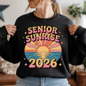 May include: Black sweatshirt with a retro design featuring the words "SENIOR SUNRISE 2026". The graphic includes a colorful sunset over water with pink, yellow, and blue hues. The design has a vintage, groovy aesthetic.