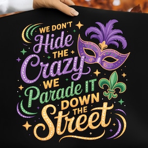 Puede incluir: Artículo decorativo negro con el texto "WE DON'T HIDE THE CRAZY WE PARADE IT DOWN THE STREET". El diseño incluye una máscara morada con plumas, un lirio verde y estrellas amarillas, verdes y moradas.