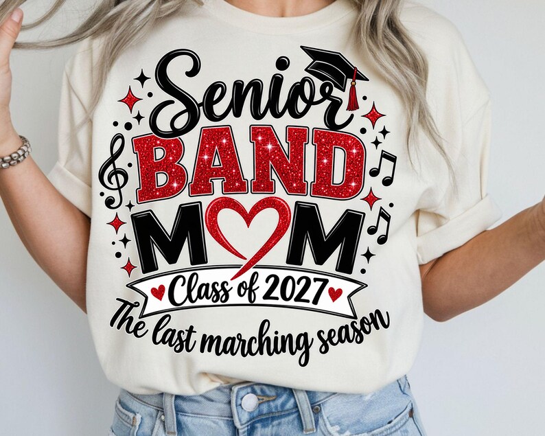 May include: A cream-colored t-shirt featuring a graphic with the text Senior BAND MOM, Class of 2027, and The last marching season. The design includes a black graduation cap, red glitter lettering, a red heart, and black musical notes.