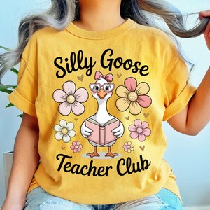 Może przedstawiać: Musztardowa ż&oacute;łta koszulka z napisem "Silly Goose Teacher Club". Koszulka przedstawia kresk&oacute;wkową gęś w okularach i kokardzie, trzymającą książkę, otoczoną r&oacute;żowymi i białymi kwiatami.