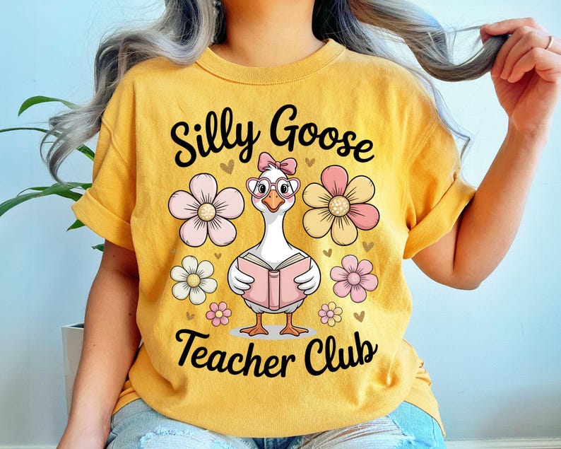 Może przedstawiać: Musztardowa ż&oacute;łta koszulka z napisem "Silly Goose Teacher Club". Koszulka przedstawia kresk&oacute;wkową gęś w okularach i kokardzie, trzymającą książkę, otoczoną r&oacute;żowymi i białymi kwiatami.