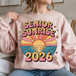 May include: A light pink sweatshirt with a colorful graphic that reads "SENIOR SUNRISE 2026." The design features a sunset over water with pink, orange, yellow, and blue hues. The text is in a retro font with a black outline.