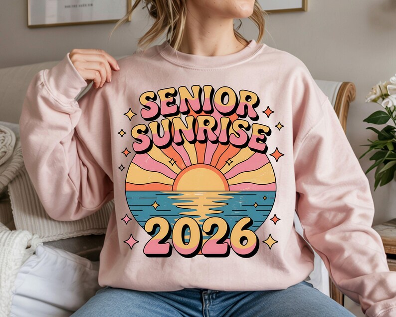 May include: A light pink sweatshirt with a colorful graphic that reads "SENIOR SUNRISE 2026." The design features a sunset over water with pink, orange, yellow, and blue hues. The text is in a retro font with a black outline.