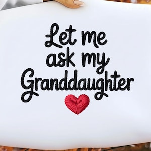 Peut inclure: Coussin blanc avec la phrase brodée "Let me ask my Granddaughter" en script noir. Un cœur rouge est brodé sous le texte. Le coussin est tenu par une personne.