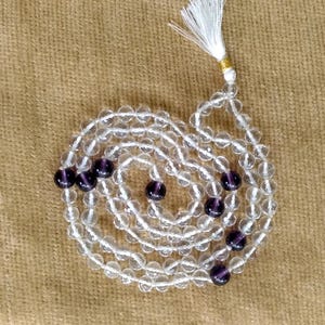 Può includere: Un mala di cristallo bianco con perline viola. Il mala è una collana di perline usata per la preghiera o la meditazione.