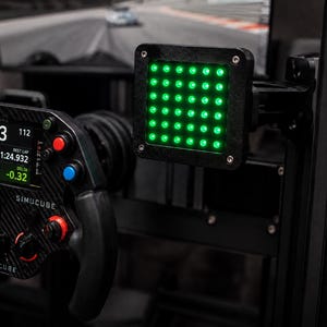 May include: A black Simucube racing steering wheel with a carbon fiber finish, featuring a digital display showing race data, red and blue buttons, and a separate square black housing containing a grid of illuminated green LED indicator lights.