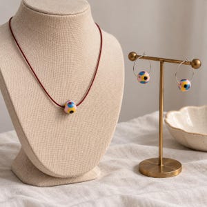 Könnte beinhalten: Eine Halskette mit einer einzelnen runden Perle mit rosa, gelben, blauen und schwarzen Punkten an einer dünnen dunkelroten Kordel, dazu passende Creolen, die jeweils eine identische Perle an einem dünnen silbernen Drahtring tragen.