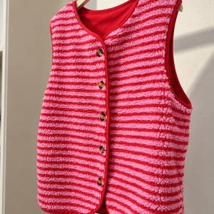 Könnte beinhalten: Eine ärmellose Weste aus strukturiertem, flauschigem Fleece-Material mit horizontalen Streifen in abwechselnd leuchtendem Pink und Rot. Das Kleidungsstück hat einen V-Ausschnitt, ein einfarbig rotes Innenfutter und eine Knopfleiste mit fünf braunen Knöpfen.