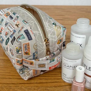May include: A rectangular, light gray cosmetic bag with a gold zipper and a handle. The bag is covered in a pattern of colorful postage stamps. Several bottles of beauty products and a bottle of nail polish are next to the bag.