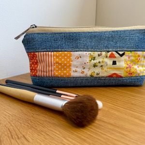 May include: A blue denim makeup bag with a patchwork design in orange, white, and yellow fabrics. The bag has a beige zip and is accompanied by makeup brushes with wooden handles and silver ferrules.