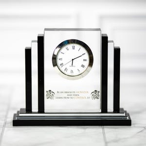 May include: A rectangular quartz clock with a silver face and Roman numerals. The clock has a black base and black vertical accents on either side. The text "BE AN ABSOLUTE MONSTER AND THEN LEARN HOW TO CONTROL IT!" is printed on the clock face.