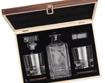 May include: A whiskey decanter set in a wooden box. The set includes a square glass decanter, two lowball glasses, a stopper, and whiskey stones. The box has a dark interior and a clasp closure.