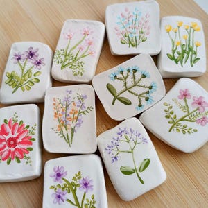 Könnte beinhalten: Eine Sammlung kleiner, quadratischer, weißer Keramikfliesen, die jeweils mit einem anderen Blumendesign handbemalt sind. Die Blumen sind in verschiedenen Farben dargestellt, darunter Rosa, Lila, Gelb und Blau, mit grünen Blättern. Die Fliesen sind auf einer Holzoberfläche angeordnet.