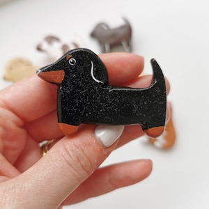 May include: A miniature black dachshund ornament with brown detailing on its snout and paws. The dog has a white curved line above its eye. Other dog ornaments are visible in the background.