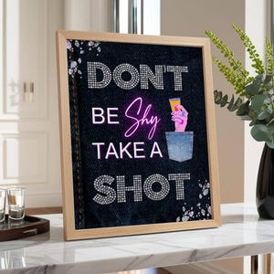 以下が含まれることがあります： ラインストーンとネオン文字で「DON'T BE Shy TAKE A SHOT」と書かれた額入りのアートワーク。アートワークはデニムの背景と木製のフレームが特徴です。ショットグラスが手前のトレイにあります。