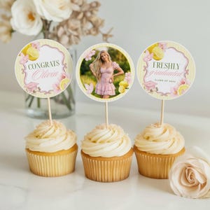 Può includere: Tre cupcake con glassa bianca, ciascuno sormontato da un topper rotondo. I topper presentano disegni a tema laurea con testo, limoni e dettagli floreali. Un topper mostra una foto.
