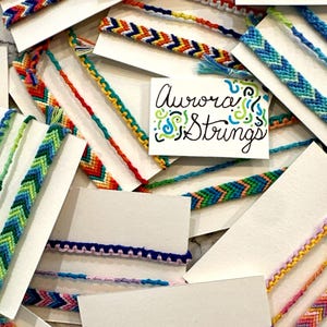 May include: A collection of colorful friendship bracelets with chevron patterns, displayed on white cards. The cards have the text "Aurora Strings" written on them.