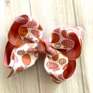 May include: A large hair bow featuring a white ribbon printed with pumpkins in shades of pink and brown. The bow has a layered design with a solid rust-colored ribbon on the underside. The bow is tied in the center with a matching pumpkin print ribbon.