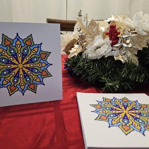 May include: Two white greeting cards with a colorful mandala design. The mandala is a circular pattern with blue, green, yellow, and orange colors. The cards are on a red tablecloth.