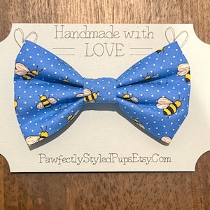 May include: A blue bow tie with white polka dots and a pattern of yellow bees. The bow tie is on a white card with the text "Handmade with LOVE" and "PawfectlyStyledPupsEtsy.com".