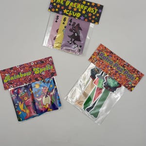 May include: Three clear plastic bags containing collectible items. One bag labeled THE BREAKFAST CLUB holds three playing cards. Another labeled Rainbow Spells contains two character cards. The third labeled Retro Pez Pals holds three candy dispensers.