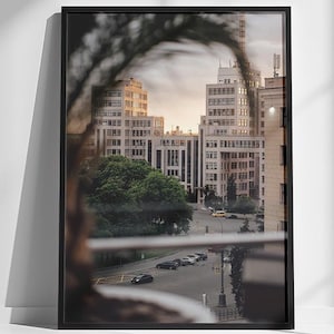 Könnte beinhalten: Ein gerahmtes Foto einer Stadtansicht mit hohen Gebäuden und einer Straße mit Autos. Das Foto befindet sich in einem schwarzen Rahmen und lehnt an einer weißen Wand. Das Bild ist gut beleuchtet, mit Fokus auf die urbane Landschaft.