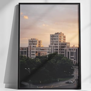 Op de afbeelding: Een ingelijste foto van een skyline bij zonsondergang. De gebouwen zijn wit en hebben veel ramen. De lucht is zacht oranje en roze.