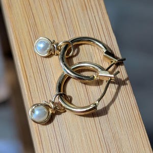 May include: A pair of gold-colored hoop earrings with a small, round, white pearl charm dangling from each hoop. The earrings are displayed on a light brown wooden surface.