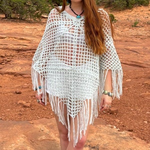 May include: A woman wearing a white crochet poncho with fringe, a white vest top, and brown cowboy boots. She is standing in front of a red rock formation.
