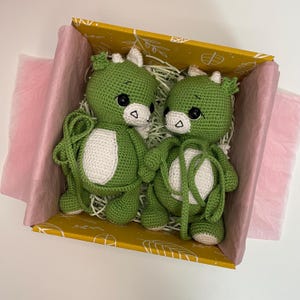 May include: Two green crocheted dragon plush toys with white bellies and horns, black eyes, and white accents, nestled in a yellow box with pink tissue paper. The dragons are holding hands, creating a charming and whimsical scene.