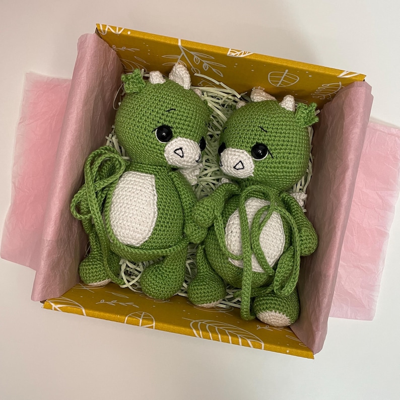 May include: Two green crocheted dragon plush toys with white bellies and horns, black eyes, and white accents, nestled in a yellow box with pink tissue paper. The dragons are holding hands, creating a charming and whimsical scene.