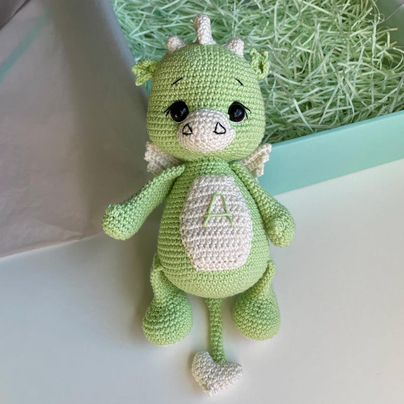 May include: A handmade green crochet dragon toy with white accents. The dragon has large black eyes, small white wings, and a white belly patch with the letter "A". The dragon's tail ends in a heart shape.