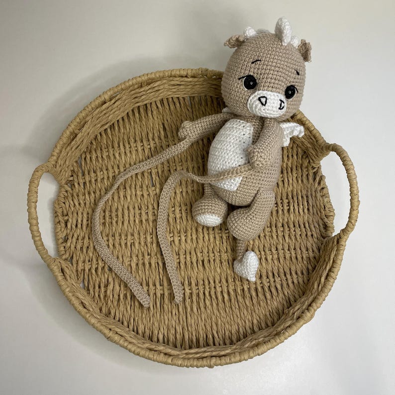 May include: A crocheted dragon doll in tan, white, and black, with long, trailing appendages and small white wings. The dragon is seated in a woven, round tray with handles. The doll has a friendly expression.