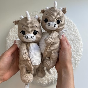 May include: Two crocheted dragon dolls in tan and white. The dragons have large black eyes, white horns, and small wings. The dolls are held in hands against a white, textured background. The dragons are a handmade craft.