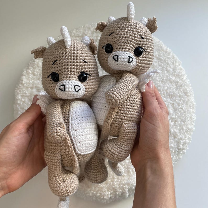 May include: Two crocheted dragon dolls in tan and white. The dragons have large black eyes, white horns, and small wings. The dolls are held in hands against a white, textured background. The dragons are a handmade craft.