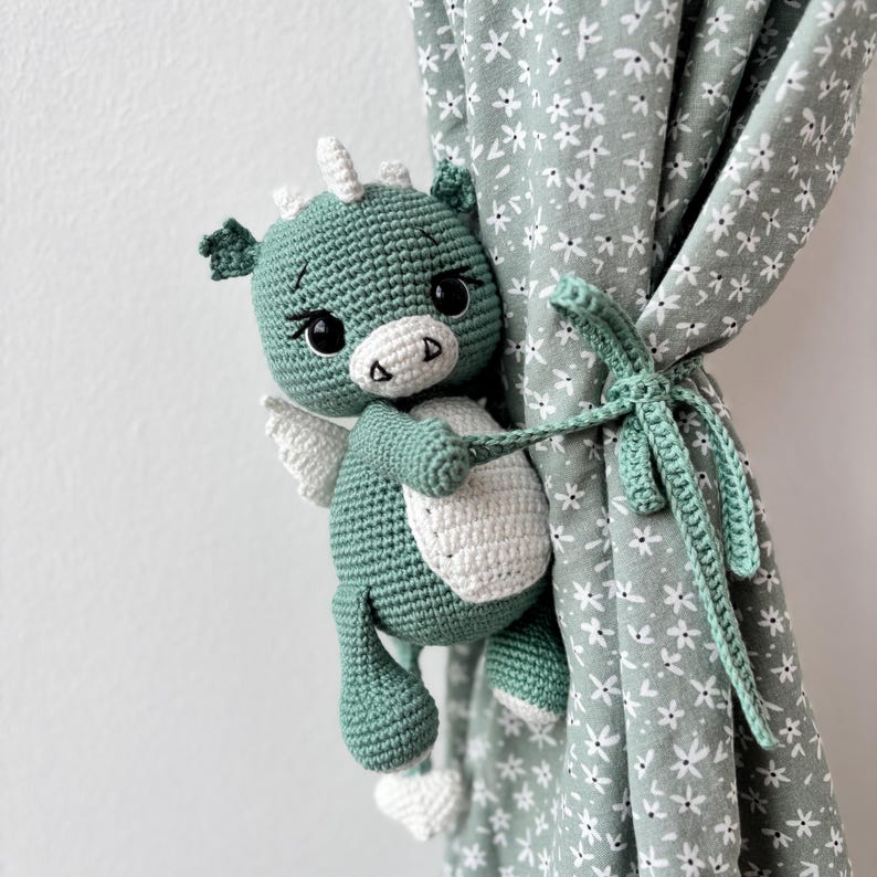 May include: A handmade crochet dragon in shades of green and white, with small wings and a friendly expression. The dragon is holding a curtain tieback, adding a whimsical touch to the room's decor. The curtain is a light green color with a white floral pattern.