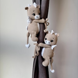 May include: Two crocheted pegasus curtain tiebacks in tan and white. Each pegasus has white wings, a white mane, and a heart-shaped tail. The tiebacks are holding onto a brown curtain.