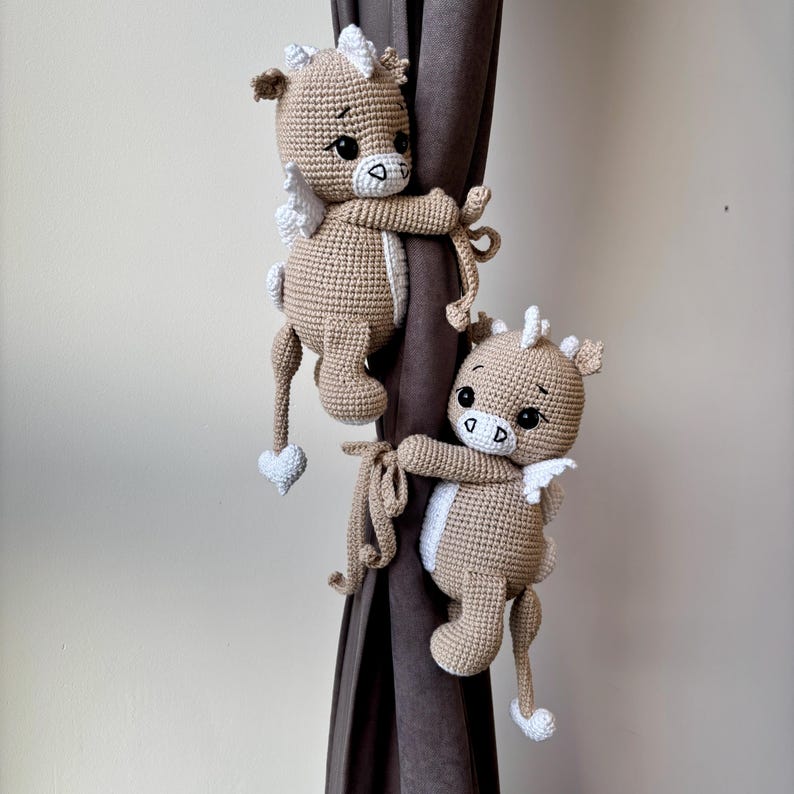 May include: Two crocheted pegasus curtain tiebacks in tan and white. Each pegasus has white wings, a white mane, and a heart-shaped tail. The tiebacks are holding onto a brown curtain.