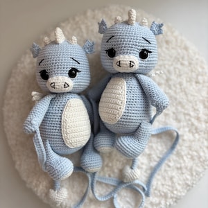 May include: Two light blue crochet dragon toys with white accents. The dragons have large black eyes, small white horns, and white wings. Each dragon has a white belly patch and is sitting on a white, fluffy surface. The dragons are handmade.