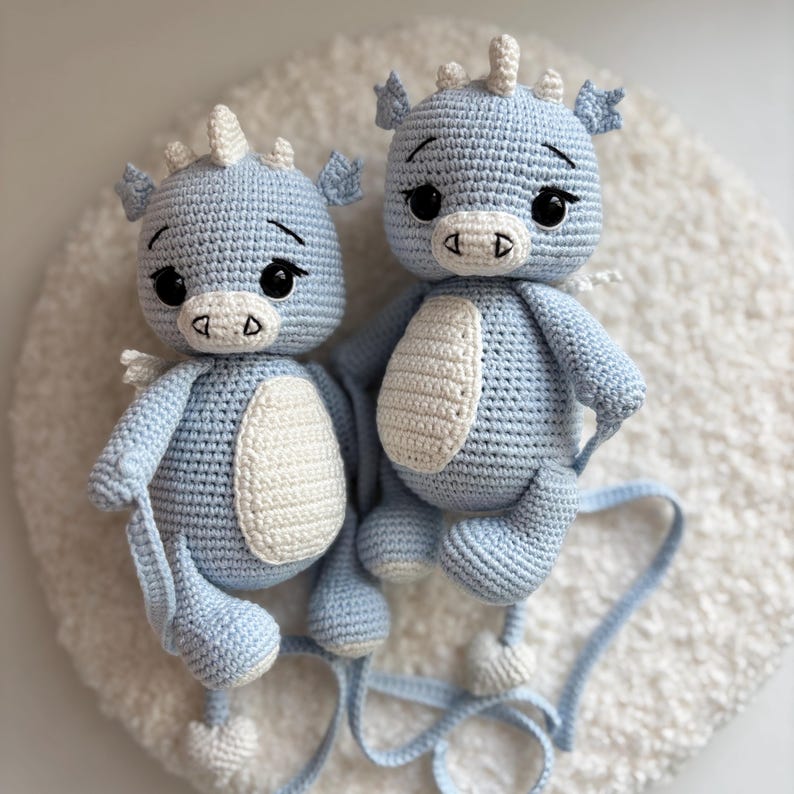May include: Two light blue crochet dragon toys with white accents. The dragons have large black eyes, small white horns, and white wings. Each dragon has a white belly patch and is sitting on a white, fluffy surface. The dragons are handmade.