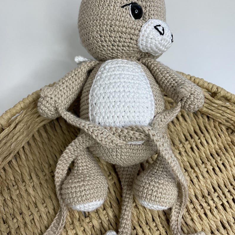 May include: A handmade crochet kangaroo doll in beige and white. The kangaroo has a white belly, black eyes, and a small black nose. The doll is sitting in a woven basket.