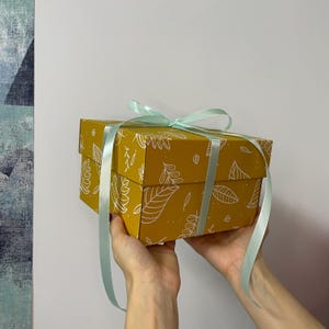 May include: A mustard-yellow gift box with a white leaf pattern, tied with a light blue ribbon. The box is rectangular and held by hands against a white background. The box is a decorative item.