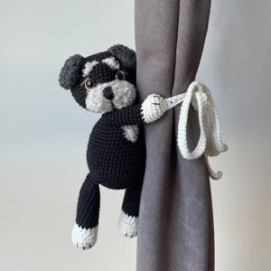 Puede incluir: Un alzapaños de cortina de ganchillo con forma de perro, con un diseño en blanco y negro y orejas texturizadas de color gris oscuro. La figura de peluche tiene ojos marrones, nariz negra y patas blancas, con largos lazos blancos tejidos.