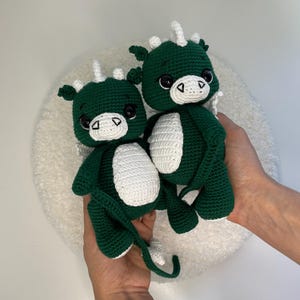 May include: Two handmade crochet dragons in dark green and white. The dragons have large black eyes, white bellies, and white horns. The dragons are being held in a person's hands. The background is a white, fluffy surface.