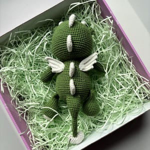 May include: A handmade green crochet dragon toy with white accents, including wings and spine details. The dragon is lying in a box filled with green paper filler, inside a pink box.