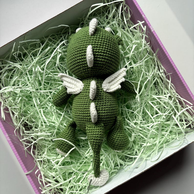 May include: A handmade green crochet dragon toy with white accents, including wings and spine details. The dragon is lying in a box filled with green paper filler, inside a pink box.