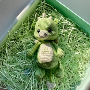 May include: A handmade green crochet dragon toy with a cream-colored belly and accents. The dragon has black eyes, small wings, and a tail. It is lying in a green gift box filled with shredded paper.