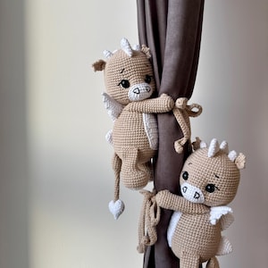 May include: Two crocheted dragon curtain tiebacks in a light brown color. Each dragon has white wings, horns, and a heart-shaped tail. They are holding onto a brown curtain, adding a whimsical touch to the room.
