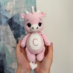 May include: A handmade pink crochet dragon doll with white accents, including wings, horns, and a heart-shaped tail. The dragon has a white oval on its chest with the letter "C". The doll is held in two hands.